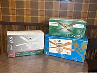Three ceiling fans in original packaging including Home Styles 42 inch white ceiling fan, Pleasantaire 42 inch ceiling fan, and Canarm 52 inch ceiling fan stacked on a table