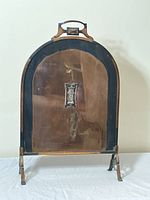 Front view of the copper and metal fireplace screen showing arched top, central decorative plate, and supporting legs.