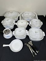 Nine Blue Cornflower Corning Ware casserole dishes of various sizes with glass lids, white Corning Ware skillet and three kitchen tools on black table