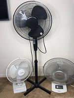 Photo showing all three fans: one black pedestal fan and two table fans, white and grey, positioned against a wall.