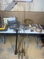 Wide view of assortment of fishing rods, tackle box with lures, fishing net, bags, and additional gear on folding table.