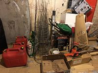 Various gardening and outdoor tools grouped in a storage area including red gas cans, wire fencing rolls, boxes, and power tool.