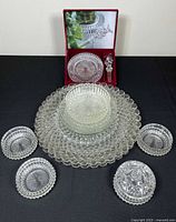 Front-facing photo showing the 2 large serving platters, 4 small plates, and 4 small bowls including one with lid, arranged on black surface, along with a boxed glass item in the back.