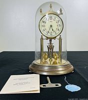 Front view of the complete clock with glass dome covering the clock body, showing the dial, rotating pendulum balls, winding key, and instruction manual on table.