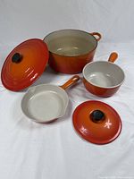 Three enameled cast iron cookware items on white backdrop: round Dutch oven with lid, saucepan with lid and 6.5″ skillet. Flame orange gradient exterior, beige interior.