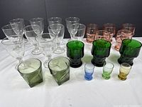Photo showing entire MCM glass lot with clear stemmed glasses, pink and green tumblers, and colorful shot glasses arranged on a white surface against a dark backdrop.