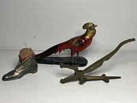 Three items: brass alligator nutcracker, metal boot pin cushion, and painted pheasant lighter placed against white background
