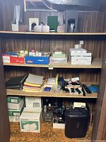 Wide view of wooden shelving unit with assorted office supplies including files, folders, small boxes, tape, and a black Staples paper shredder on bottom shelf