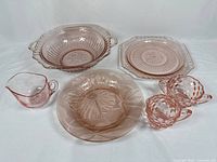 Overall view of six pink glassware items including bowl, plates, sugar, creamer, and small pitcher arranged on white background.
