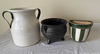 Room lit image showing all three pottery items placed side by side on a white cloth surface: the white crackled pot with metal handles, black three-legged pot, and green-white ceramic wall hanger.