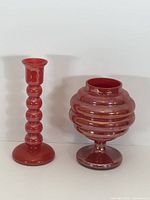 Photo of two items side-by-side on white surface: cherry red candlestick holder and iridescent red glass vase with ribbed design.