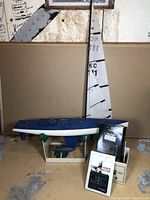 Vintage remote control sail boat with its large sail upright beside it, radio controller, and original packaging box arranged on floor.