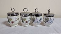 Front view showing four Royal Worcester egg coddlers with different floral and berry designs on porcelain bodies and metal looped lids.