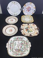 Seven decorative serving platters of various sizes and patterns arranged on black background: two large round with handles, three smaller plates with floral motifs, two square decorative platters with handles and detailed floral/bird designs.