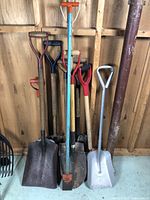 Photo showing assortment of eight shovels standing against a wooden wall, variety in blade shape, handle material, and size evident.