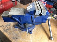 Side view of blue metal vise with swivel base, showing serrated jaw and mounting holes on base.