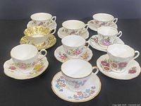 Full view of nine assorted porcelain teacups with matching saucers showing floral and bird designs with gold accents.