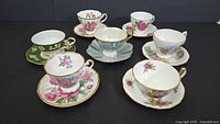 Seven assorted vintage teacups and saucers arranged on black surface showing floral and gold trim details.