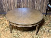 Front angle view of a vintage round wooden coffee table on brown patterned carpet against wood-paneled wall.