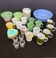 Group shot of teal blue mixing bowl at back right, two pale green bowls at back left, stack of nine white opal glass bowls with pastel stripes top center, four olive green pedestal mugs front right
