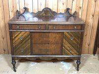 Full front view of vintage wooden sideboard showing decorative top crest, three upper drawers with brass knobs and lower paneled doors with brass handles and inlaid wood design