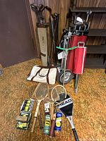 Vintage golf clubs with beige and red bags and push cart, Adidas sports bag, wooden tennis rackets, and boxes of balls on carpet