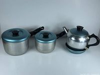 Set of two aluminum pots with aqua blue lids and black handles next to aluminum tea pot with aqua blue lid and base on white background