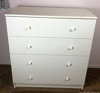 White dresser with four drawers and two simple round knobs per drawer, set against a pale pink wall and carpeted floor.