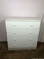 White four drawer dresser shown from the front in a room with carpet flooring and white walls. Light wear visible consistent with usage.