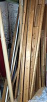 Photo showing vertical stacked wood planks including 2x4 lumber and trim pieces, with visible wood grain and some nails or screws marks, a beige painted trim piece, and some metal unfinished metal corner protectors nearby.