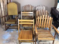 View of all wooden chairs in lot showing rocking chair, flat back chairs, and slat back chairs grouped together on concrete floor.