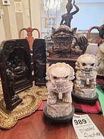 View showing front of the two white plaster Foo Dogs, seated Buddha figurine in carved niche, and two dark brown resin standing Asian figures on a table.