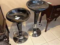 Pair of black adjustable bar stools with chrome metal bases, positioned side by side on a tiled floor next to wooden furniture.
