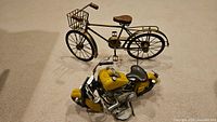 Photo showing both the decorative bicycle and the yellow motorcycle figurine together on a carpeted floor.