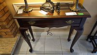 Front view of console table showing glass inset top, single drawer with brass pull and curved legs