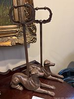 Pair of brown metal dog-shaped boot jacks with vertical handles and decorative grips, placed on a wooden surface near a framed painting.