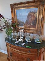 Photo showing two lantern style candle holders with metal handles, vase with faux flowers, and two glass terrariums with faux succulents and white stones, arranged on a dark green marble top wooden dresser under a framed painting.