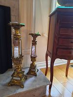 Two gold-finished ornate candle holders with mirrored panels on a marble surface next to wooden furniture showing overall lot contents.