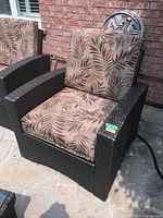 Front view of one dark brown faux wicker patio chair with leaf-patterned upholstered removable cushions placed on a stone patio against a brick wall.