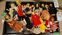 Top-down photo showing 12 collector dolls arranged on a black surface, dressed in traditional costumes from various cultures, some with small damages.