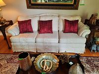Full front view of white patterned upholstered couch with three red pillows on a hardwood floor next to side tables and rug.