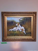 Full view of the framed painting showing two dogs lying on grass with a fence and foliage background in an ornate gold frame.