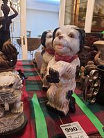 Two ceramic dogs standing upright with black patches, white fur, and red bows, placed on a checkered cloth surface with various other decorative items in the background.