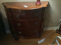 Front view of three-drawer wood dresser with curved front, ornate drawer pulls, and side shelving.