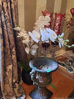 White orchid faux flowers in black pot alongside decorative urn pot with sculpted handles and detailed base, medium wear inside urn.