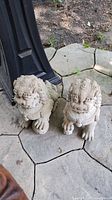 Two weathered cement Foo Dogs placed outdoors on stone pavers with dirt and foliage in the background.