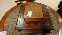 Two wooden game boxes stacked on a round table, one smaller lighter-colored box on top and a larger darker checkerboard box below.