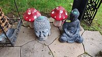 Four garden decorations on a patio: two large red mushrooms with white heart spots, one Buddha head statue, and one seated Buddha figure statue.