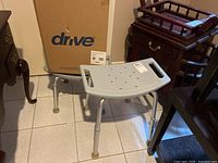 Separate gray plastic shower seat with perforations, adjustable aluminum legs with brown rubber tips, and Drive brand boxed toilet seat riser in background.