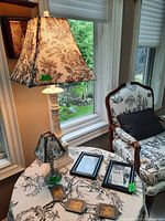 Photo showing large white lamp with toile-patterned shade, small silver lamp with shade, three metal frames lying flat on table, and floral patterned chair